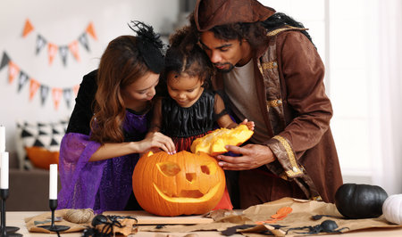 Young african american family in Halloween costumes mother father and cute little girl daughter making together jack o lantern from pumpkin, parents with kid preparing for celebration at homeの写真素材