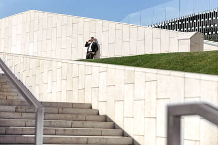 Young satisfied businessman in formal suit speaking on mobile phone smartphone while standing on concrete stairs outside during coffee break at work, office worker enjoying sunny day outsideの写真素材