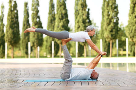 Mature retired family husband and wife practicing advanced couple yoga position on mat in city park on sunny morning, senior man holding his female partner woman on outstretched legsの写真素材