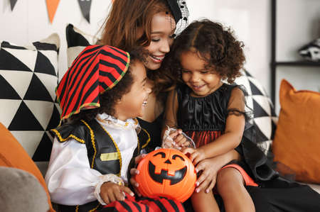 Happy african family smiling mother and two kids celebrating Halloween holiday together at home, mom with son and daughter playing with pumpkin lantern while sitting together on sofa and smilingの写真素材