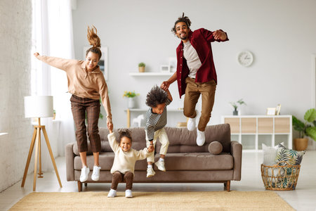 Active young african american parents jumping from sofa to floor with two little kids at homeの写真素材