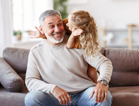 Cute little girl covering eyes with hands of her smiling grandfather while playing together at homeの写真素材