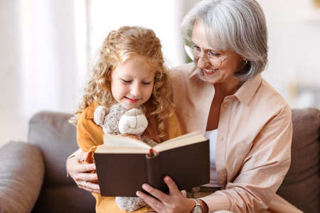 Cute little girl granddaughter reading book with positive senior grandmotherの写真素材