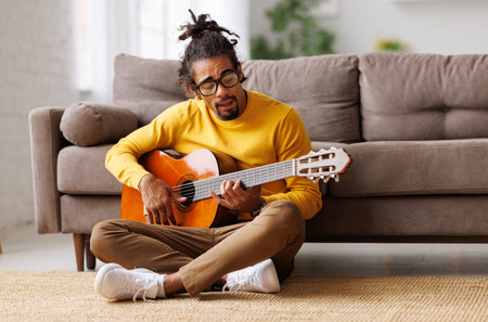 Young joyful african american man playing acoustic guitar at home, sitting on floor in living roomの写真素材
