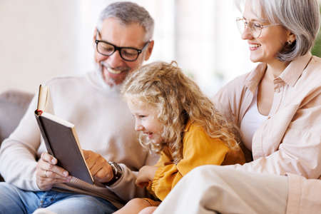 Cute little girl granddaughter smiling when reading book with senior grandparentsの写真素材