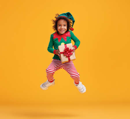 Happy african american kid boy Christmas elf costume with wrapped Xmas gift in hands jumping in airの写真素材