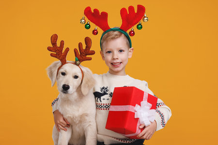 Happy preschool boy with Christmas gift hugging Golden Retriever puppy, wearing red reindeer hornsの写真素材