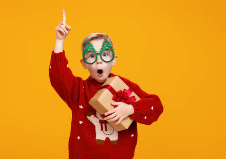 Smiling little boy wearing funny glasses in form of Christmas trees pointing with finger upside holding Xmas giftの写真素材