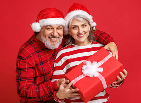 cheerful senior family couple dressed in red Santa hats holds a Christmas gifts and laughing a colored red backgroundの写真素材
