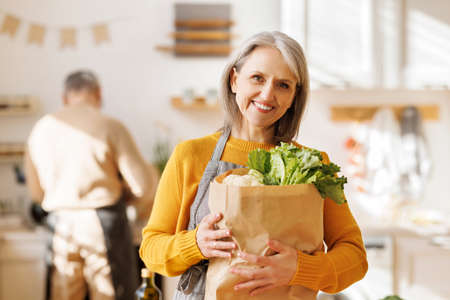 happy elderly woman holds a bag of vegetables in her hands at home in the kitchenの写真素材