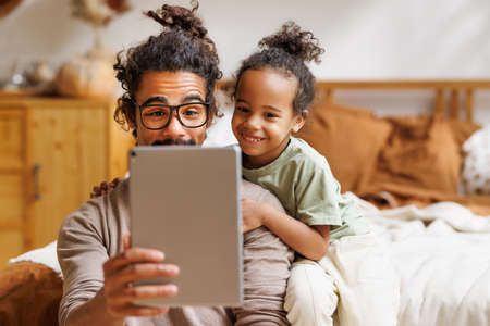 Happy african american family little son with dad watching funny videos on tablet together at homeの写真素材