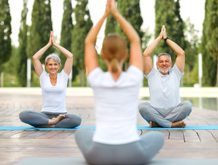 Senior couple staying in lotus pose in front of female yoga trainer during yoga class in city parkの写真素材