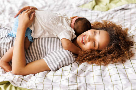 happy african american mother gently hugs baby daughter in bedの写真素材
