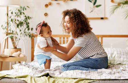 happy african american mother playing with laughing baby daughter in bedの写真素材