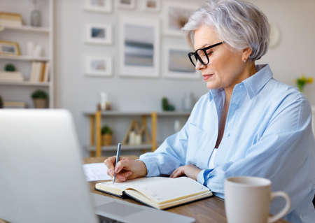 Mature businesswoman working at laptop, reading documents and take notes in home officeの写真素材