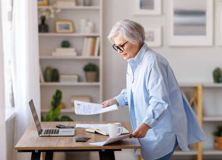 Mature businesswoman working at laptop, reading documents and take notes in home officeの写真素材