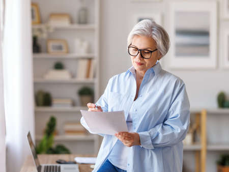 Mature businesswoman working at laptop, reading documents and take notes in home officeの写真素材