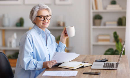 Mature happy businesswoman working at laptop, reading documents and take notes in home officeの写真素材