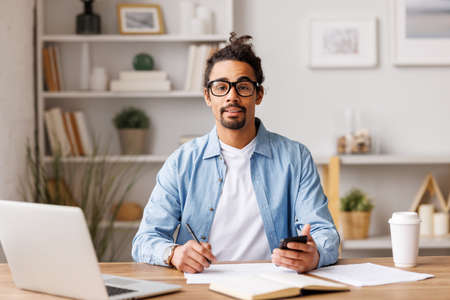 Young african american man freelancer working remotely on laptop and browsing smartphone or studying online at homeの写真素材