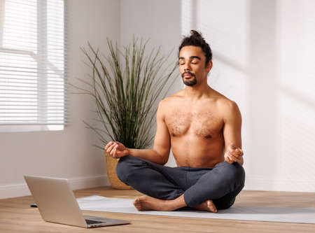 Relaxed black man meditating in lotus pose in front of laptop at homeの写真素材