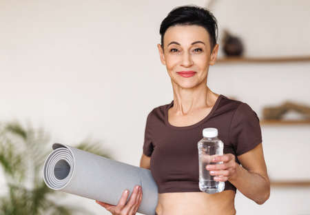 Mature sportswoman with mat and water at homeの写真素材