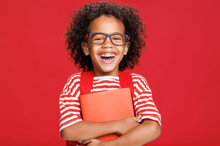 Happy ethnic schoolboy laughing with book against red backgroundの写真素材