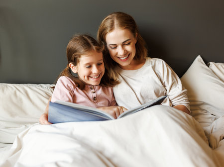 Happy mother and daughter reading book together on bedの写真素材