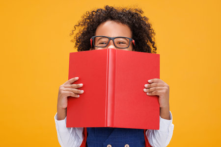 Happy sly curly schoolboy looks out from behind the book