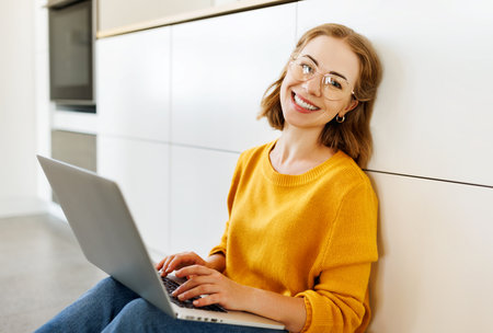 cheerful young woman uses a laptop at home sitting on floor in a modern kitchenの写真素材