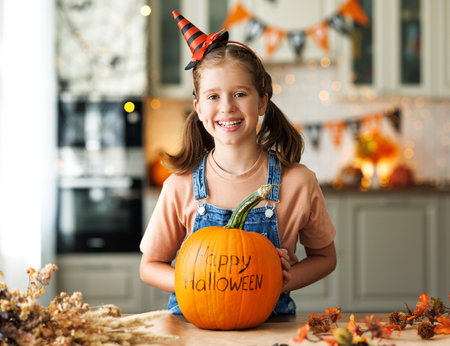 Smiling kid girl in hat showing pumpkin with inscription Happy halloween during Halloween celebrationの写真素材