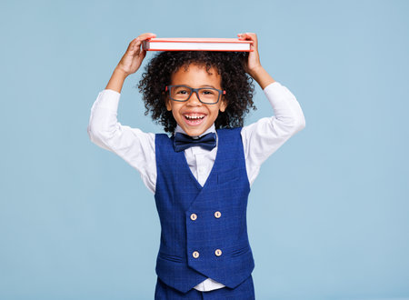 Happy ethnic schoolboy playing with book against blue backgroundの写真素材