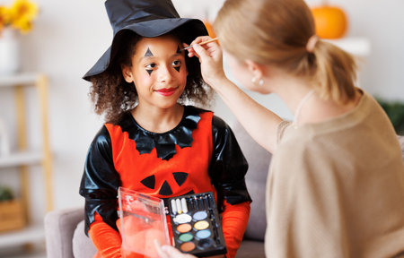 Festive makeup for Halloween. Woman doing witch make-up for ethnic curly girl in costume while preparing holidayの写真素材