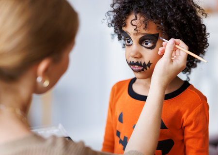 Festive makeup for Halloween. Woman doing pumpkin make-up for ethnic curly boy in costume while preparing holidayの写真素材