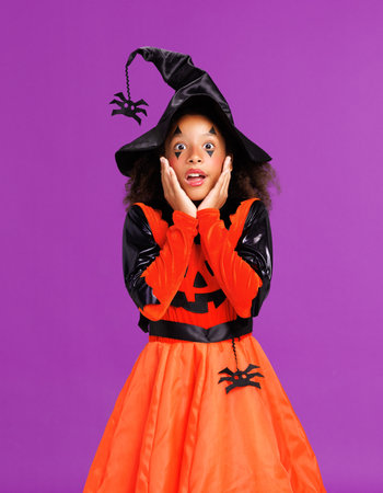 Shocked cheerful ethnic girl in costume pumpkin touches cheeks in surprisewhile celebrates Halloweenの写真素材