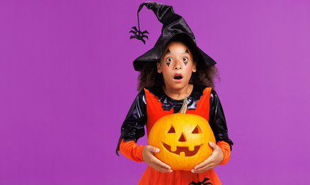 Happy ethnic girl in costume holding pumpkin and screams in surprise while celebrates Halloween on purple backgroundの写真素材