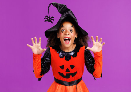 Happy ethnic girl in a pumpkin costume scary gestures and celebrates Halloween and laughs on bright purple backgroundの写真素材