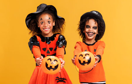 Happy ethnic children in pumpkin costume with basket celebrates Halloween and laughs on yellow backgroundの写真素材