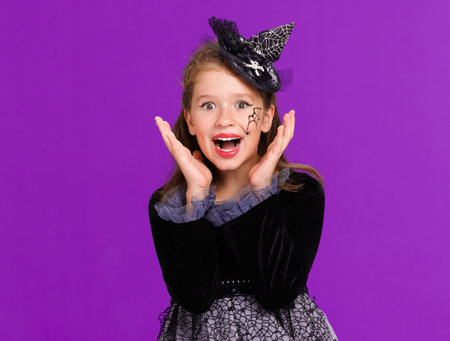 Shocked cheerful girl in costume witch touches cheeks in surprise while celebrates Halloween on purple backgroundの写真素材