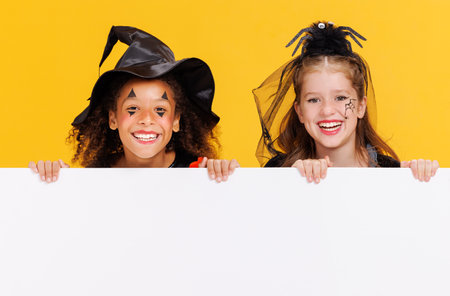Happy Halloween. Children girls in carnival costumes and makeup with blank white poster on colored yellow backgroundの写真素材