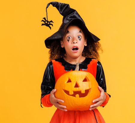 Happy cheerful african american girl with curly hair in costume holding pumpkin and screamsw in surprise while celebrates Halloween on yellow backgroundの写真素材