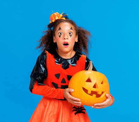 Happy cheerful african american girl with curly hair in costume holding pumpkin and screamsw in surprise while celebrates Halloween on blue backgroundの写真素材