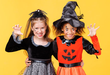 Two happy cheerful kids girls in witch costume and terrible makeup scary gestures and celebrates Halloween and laughs on yellow backgroundの写真素材