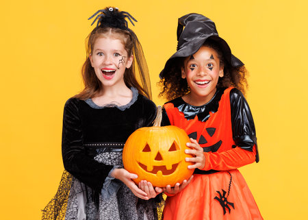 Two happy cheerful kids girls in witch costume and terrible makeup with pumpkin jack-o-lantern celebrate Halloween and laughs on yellow backgroundの写真素材