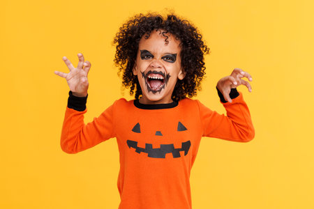 Happy cheerful african american boy in pumpkin costume and terrible makeup make scary gestures and celebrates Halloween and laughs on yellow backgroundの写真素材