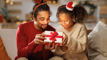 Happy afro american curly girl with father opening christmas present, excited kid daughter with loving dad on couch. New Year traditions for familiesの写真素材