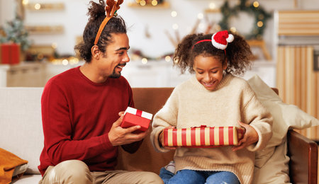 Happy afro american curly girl with father exchange christmas present, excited kid daughter with loving dad on couch. New Years traditions for familiesの写真素材