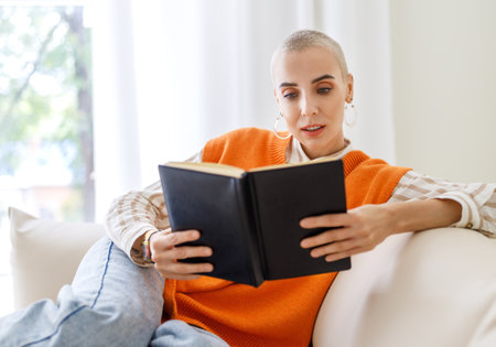 Beautiful woman with very short hair reading book while sitting on sofa at home in daytimeの写真素材