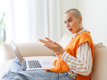 Beautiful woman freelancer with short hair works, makes video call and surprised gestures while sitting on sofa and working on laptop during work from homeの写真素材