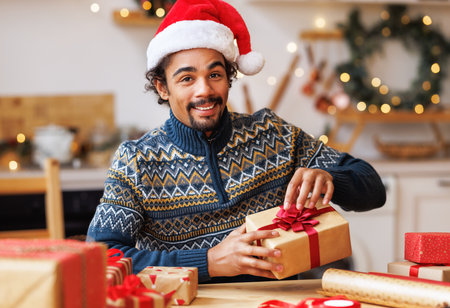 Delighted african american man smiling and wrapping decorative paper around present box while sitting at table and preparing for Christmas celebration at homeの写真素材