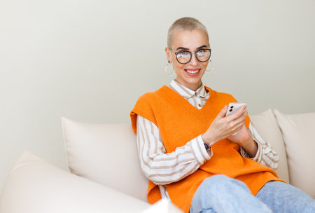 Young beautiful woman with very short hair sitting on sofa and using smartphone, texting message or surfing internet while relaxing at home. female with cell phoneの写真素材